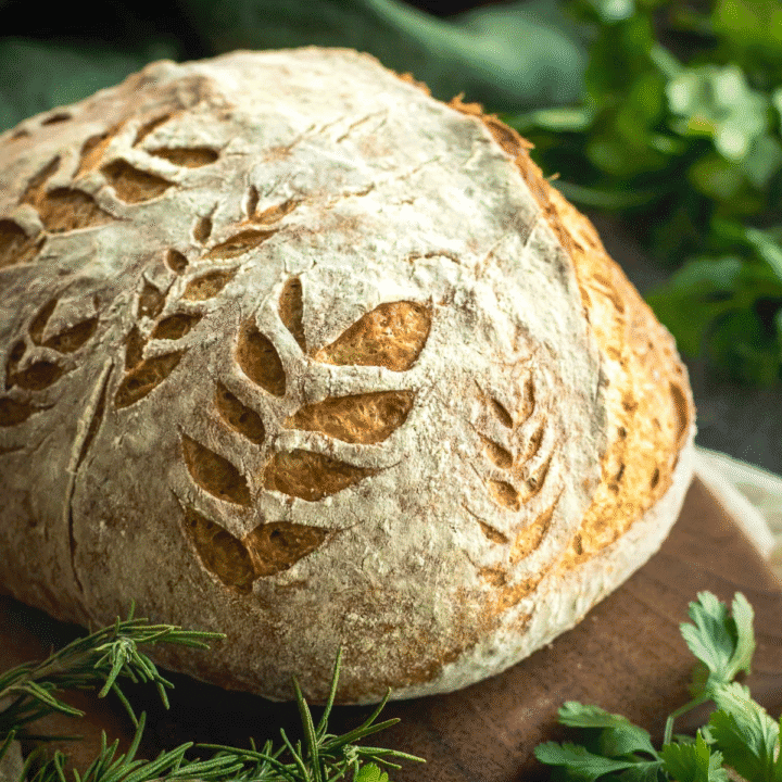 A loaf of rustic bread on a wooden board dusted with flour and scored with leaves.