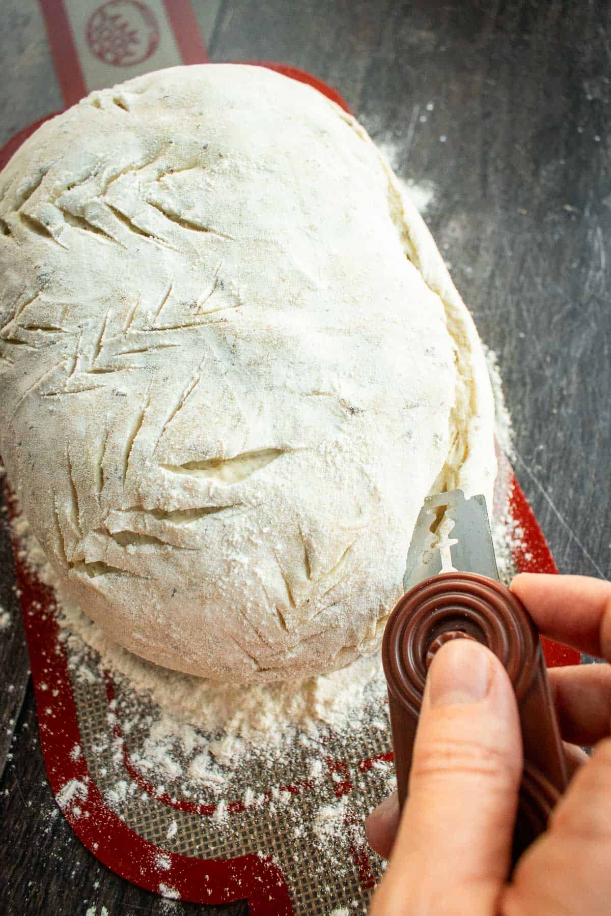A hand scoring a loaf of bread dough with a bread lame.