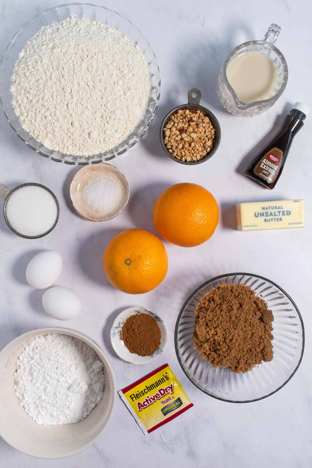 Oranges and a stick of butter next to bowls of flour, brown sugar, milk, salt, white sugar, and chopped nuts on a light surface.