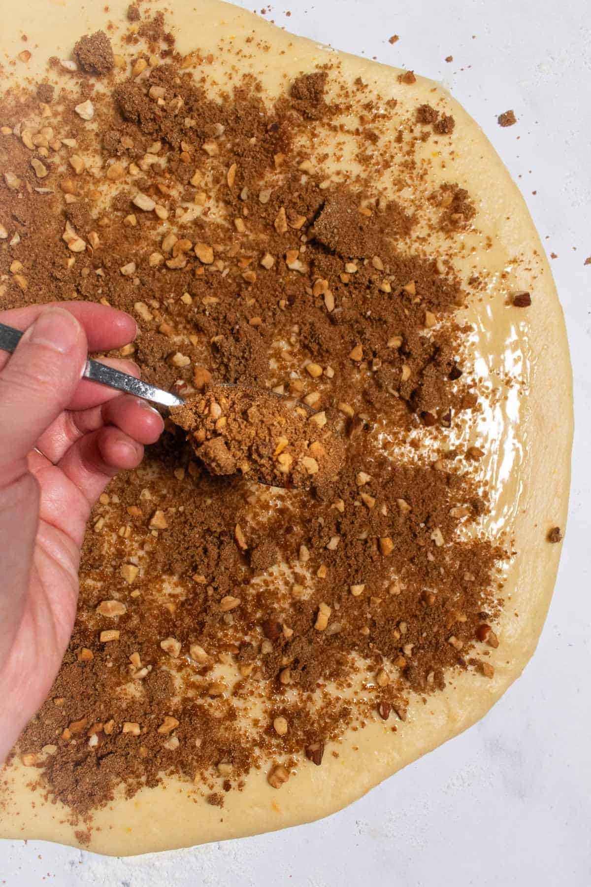 A hand sprinkling cinnamon sugar and chopped nuts over rolled-out bread dough.