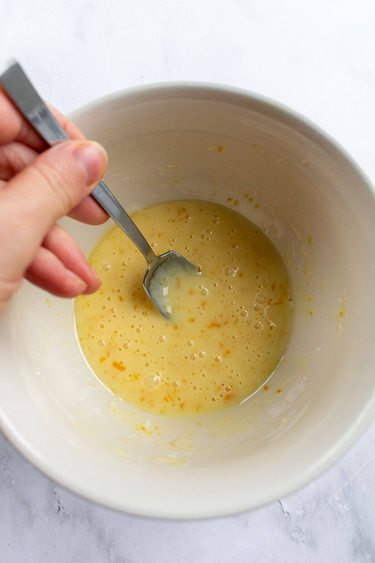 A hand mixing orange glaze in a bowl with a spoon.