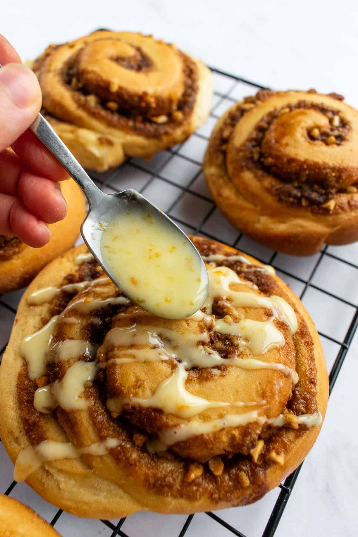 A hand spooning orange glaze onto baked orange cinnamon rolls.