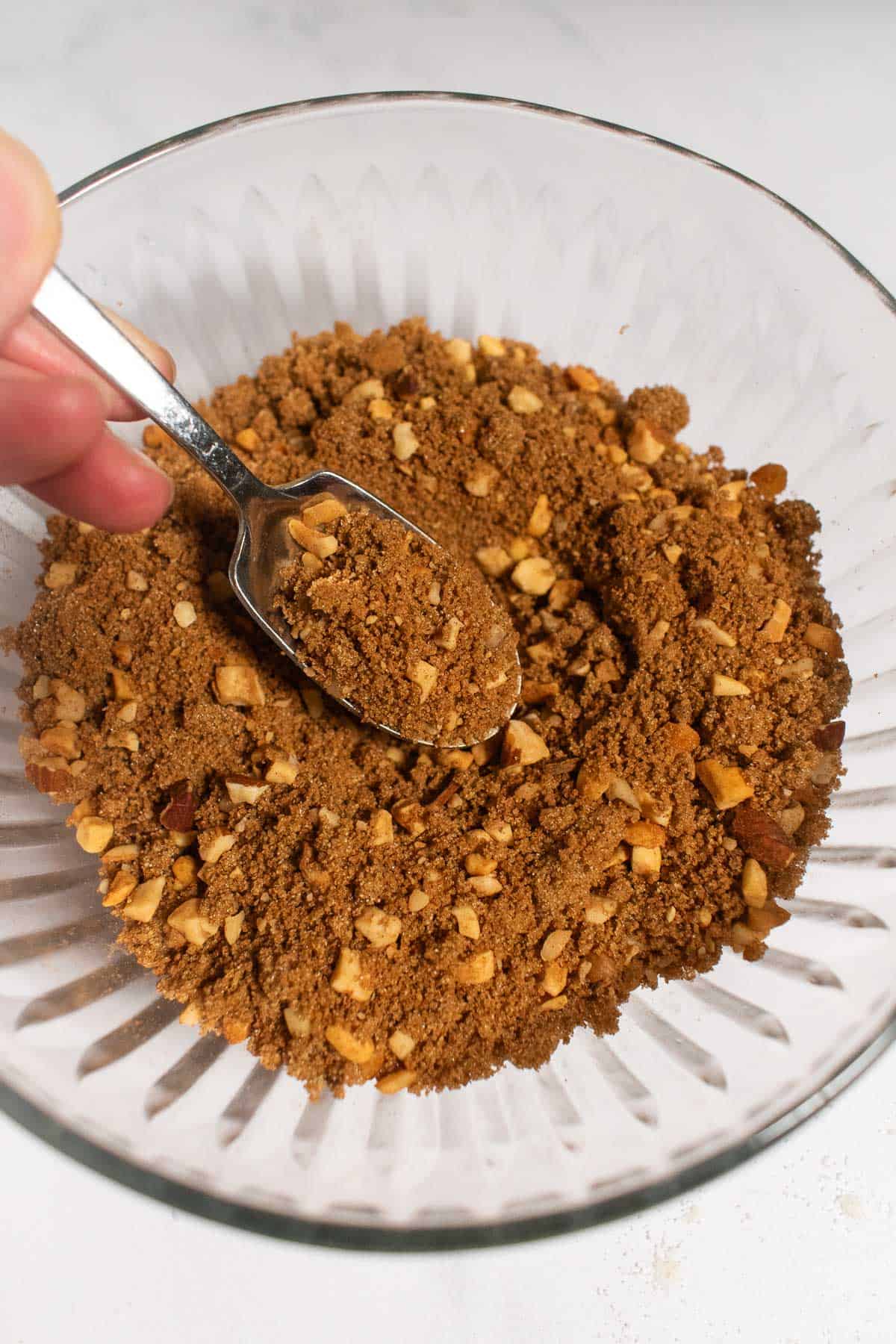 A hand mixing cinnamon, sugar, and chopped nuts in a glass bowl.