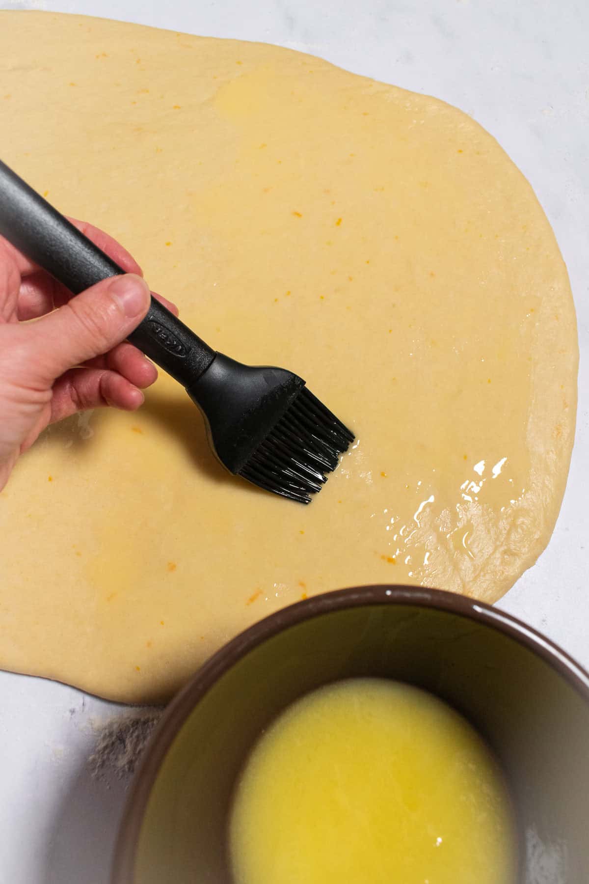 A hand brushing melted butter over rolled-out bread dough.