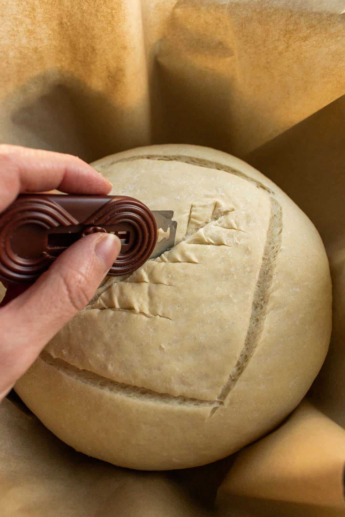 A hand using a bread lame to score dough inside a Dutch oven.