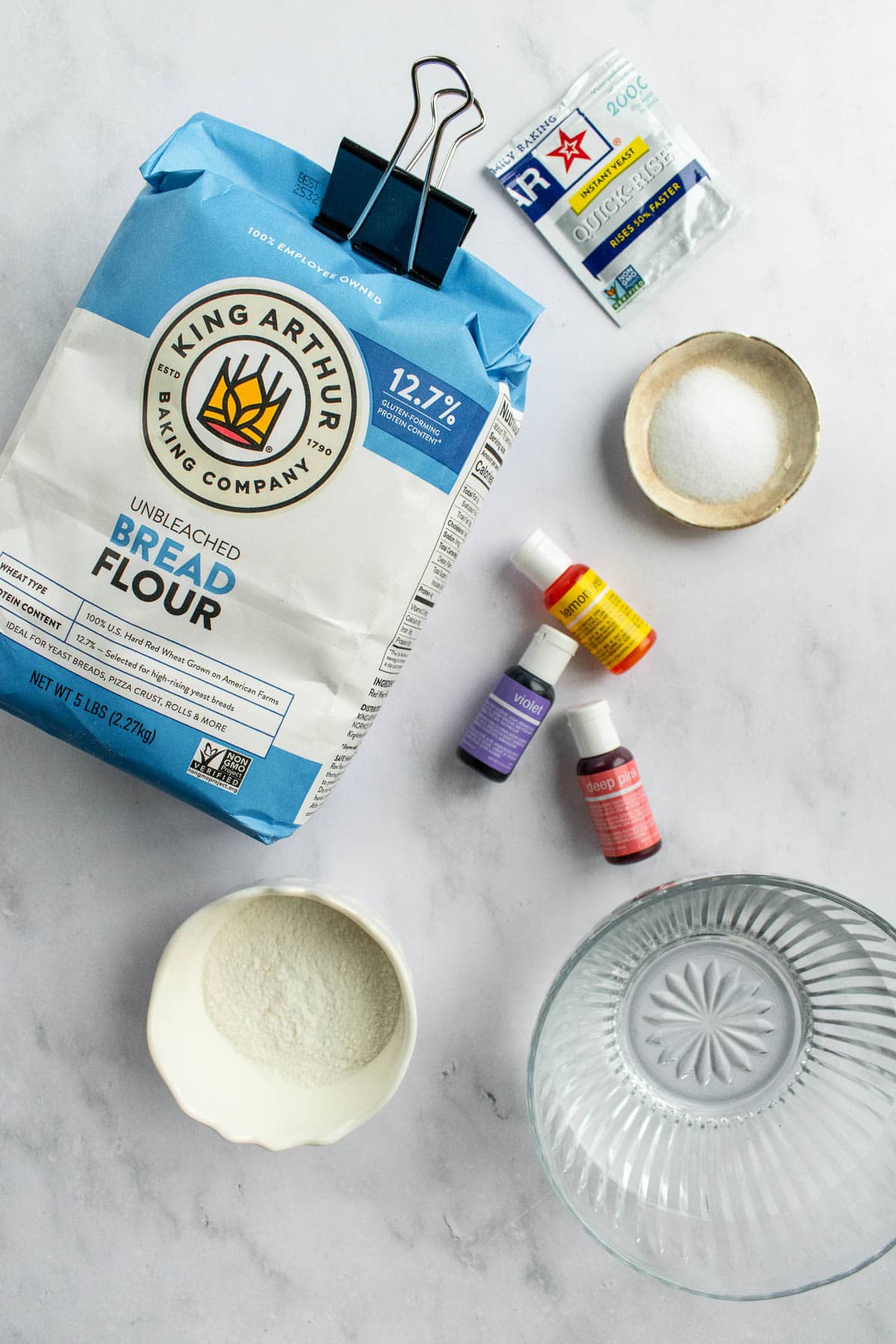 A bag of bread flour next to bowls of water, salt, and rice flower, a packet of yeast, and bottles of food dye on a marble surface.