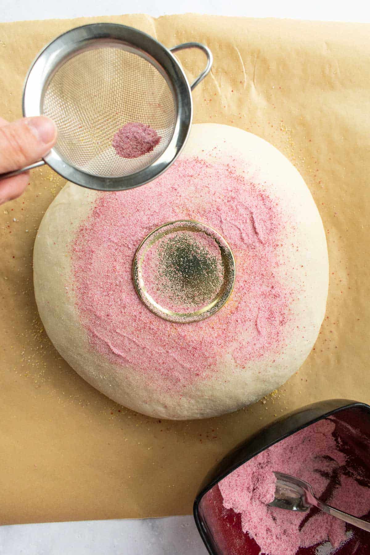 A hand sifting pink rice flour on a boule of bread dough.