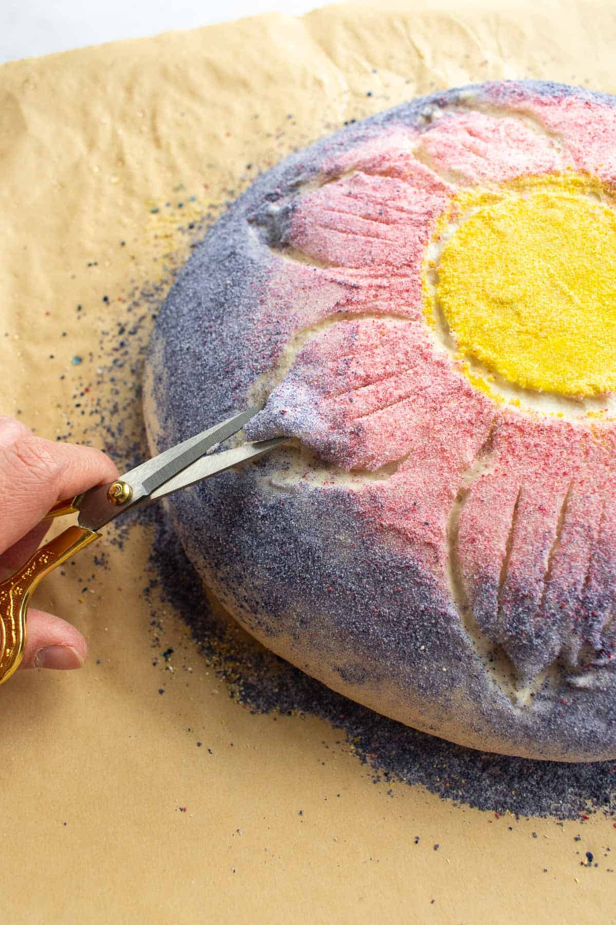 A hand using scissors to cut the tips of flower petals as a design into a ball of bread dough that's dyed purple and pink.