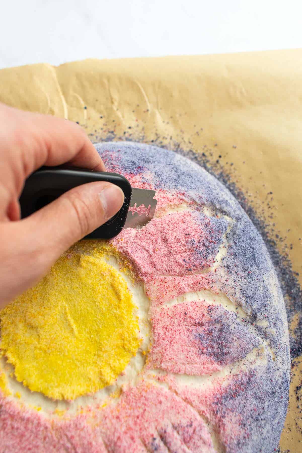 A hand scoring flower petals into pink and purple-dyed bread dough with a bread lame.