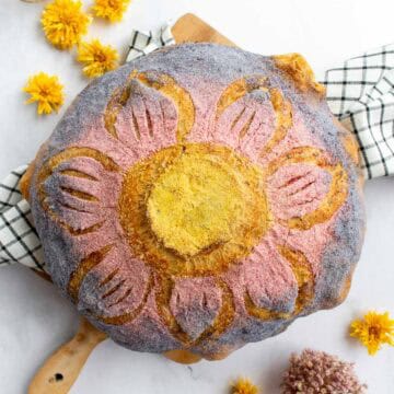 A loaf of bread scored like a flower and dyed with yellow, pink, and purple powder on top.
