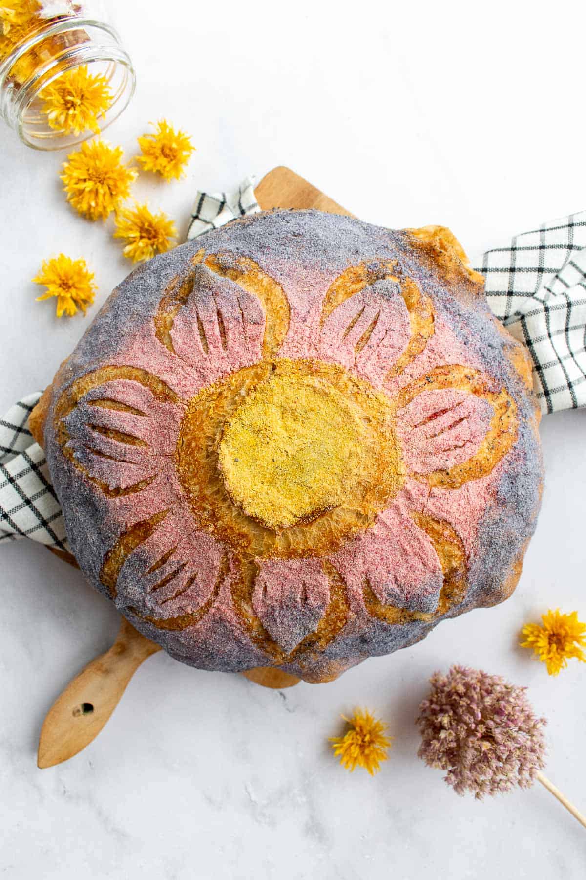 A loaf of bread scored like a flower and dyed with yellow, pink, and purple powder on top. 