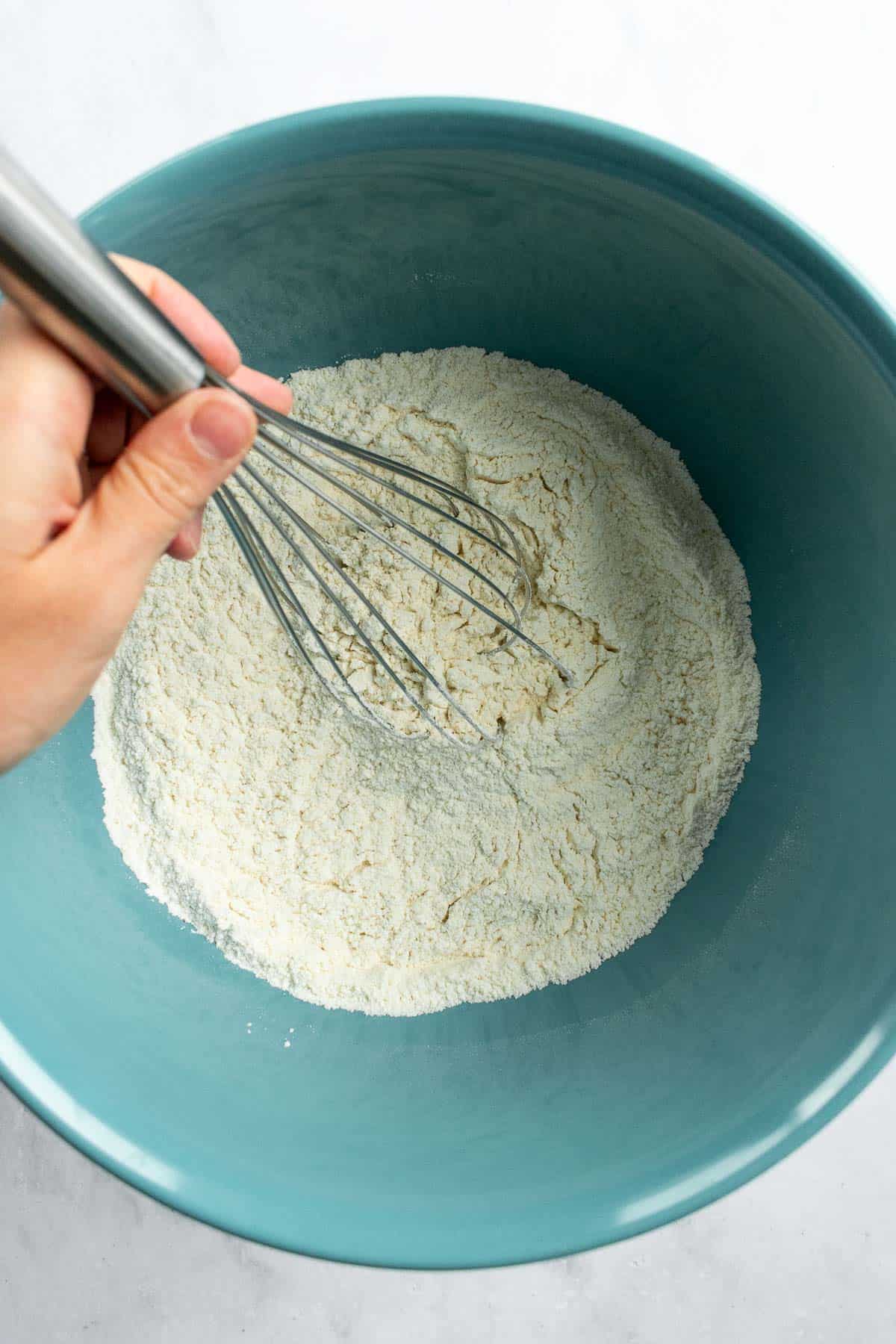 A hand whisking flour, salt, and yeast in a large blue bowl.