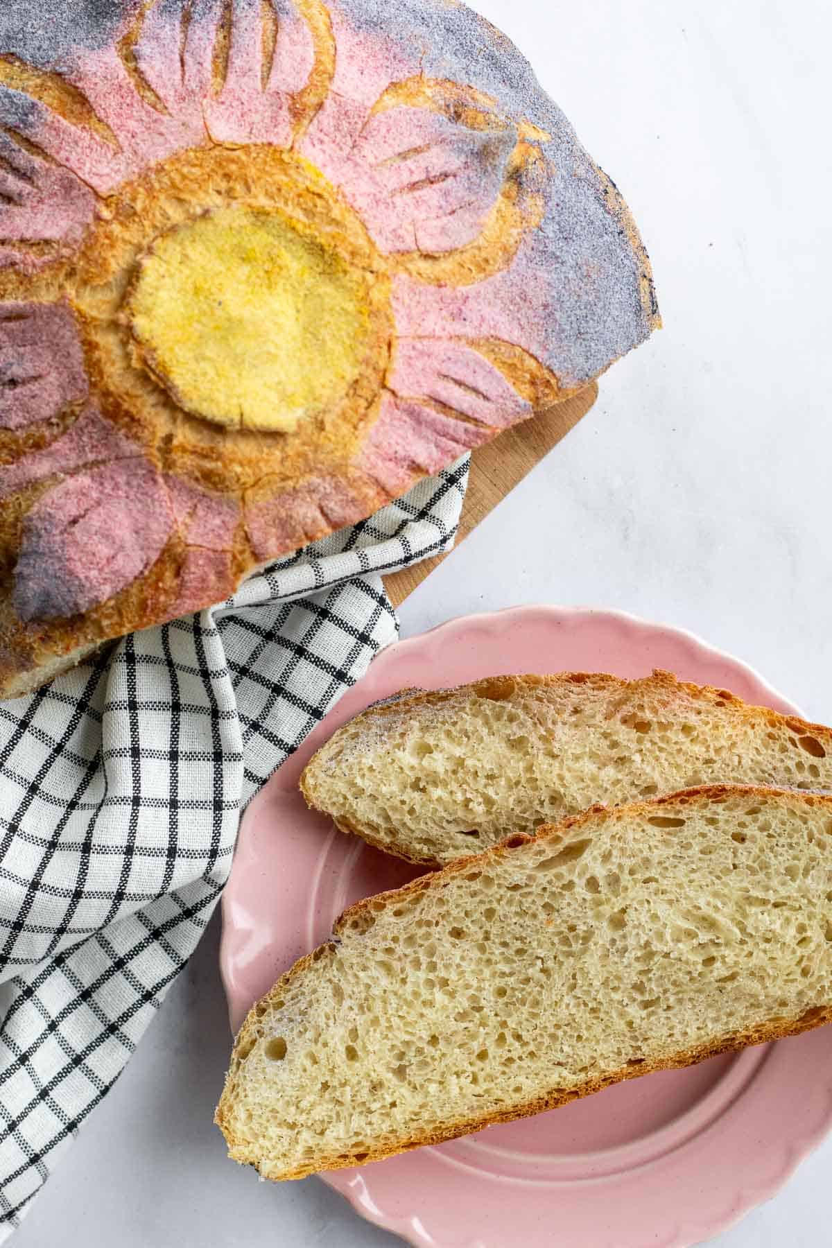 Two slices of rustic white bread on a pink plate next to a loaf of bread dyed and scored like a flower.