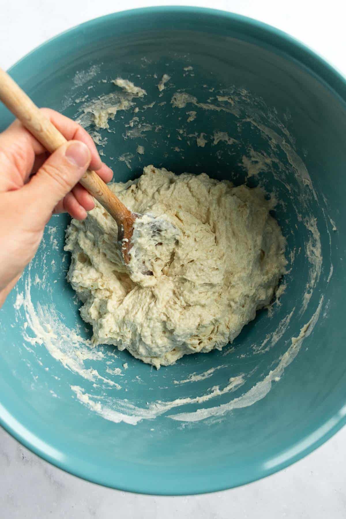 A hand mixing white bread dough in a bowl with a wooden spoon.