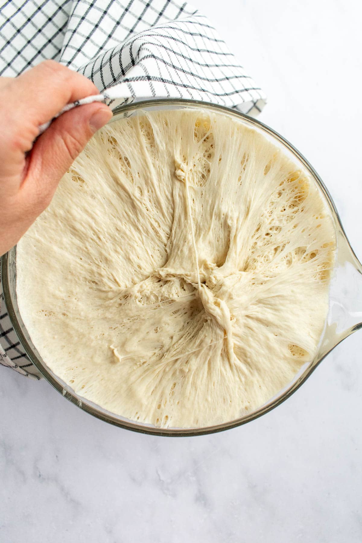 Risen, bubbly bread dough in a glass mixing bowl partially covered by a hand holding a linen.