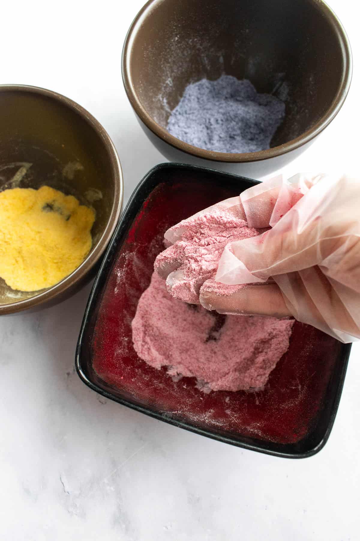 A gloved hand mixing a pink powder in a bowl next to bowls of yellow and purple powder.
