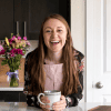 A girl with a pink apron and a mug leaning against a kitchen counter.