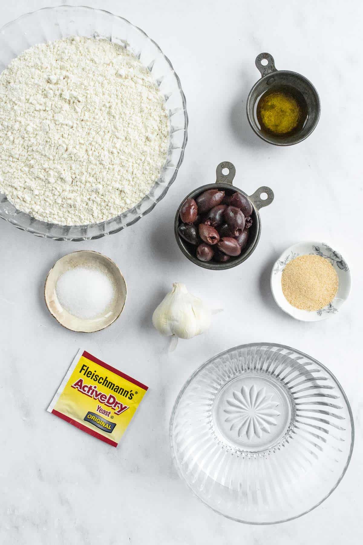 Bowls of water, flour, Kalamata olives, olive oil, garlic powder, and salt next to dry yeast and a head of garlic on a table.