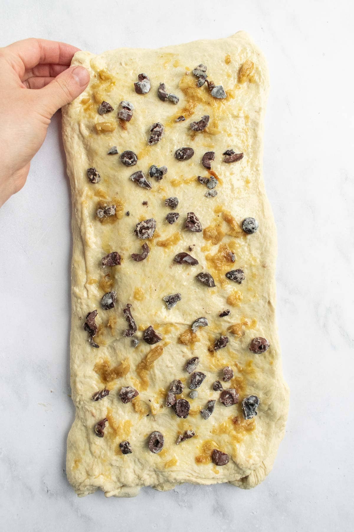 A hand holding a corner of a large rectangle of dough covered in mashed garlic and chopped olives.