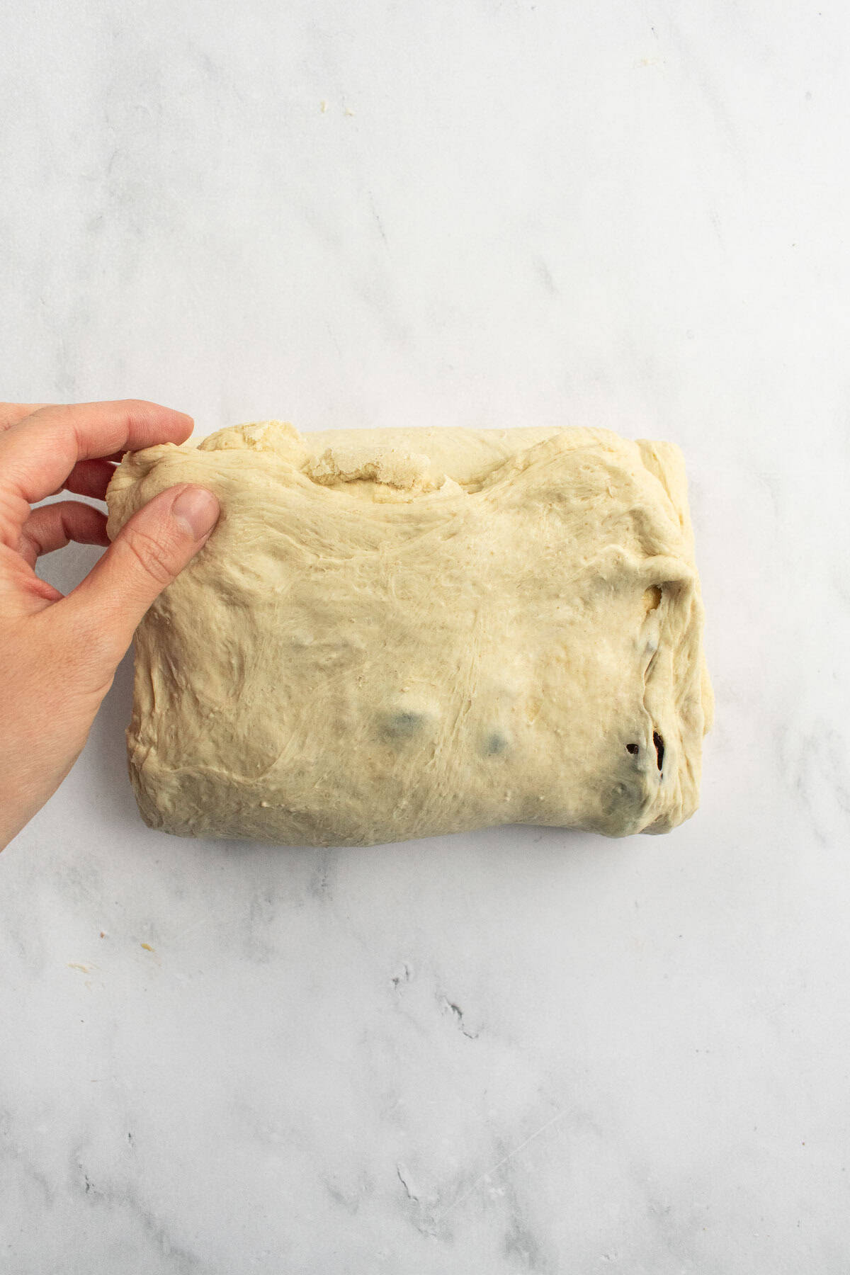 A hand holding a corner of a rectangle of olive bread dough on a marble surface.