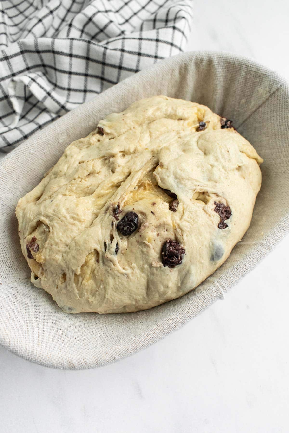 A loaf of olive bread in a proofing basket on a light surface.