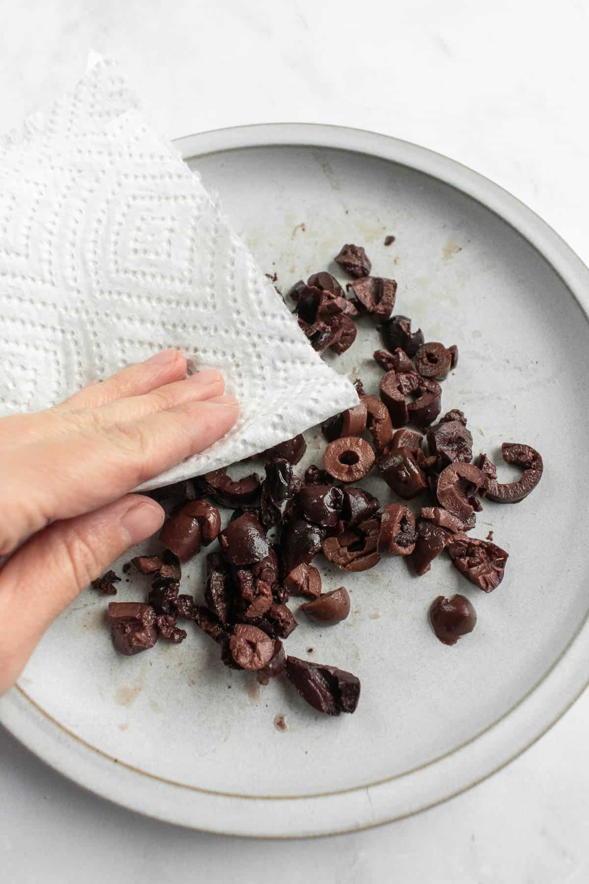 A hand patting chopped olives on a plate with a paper towel.