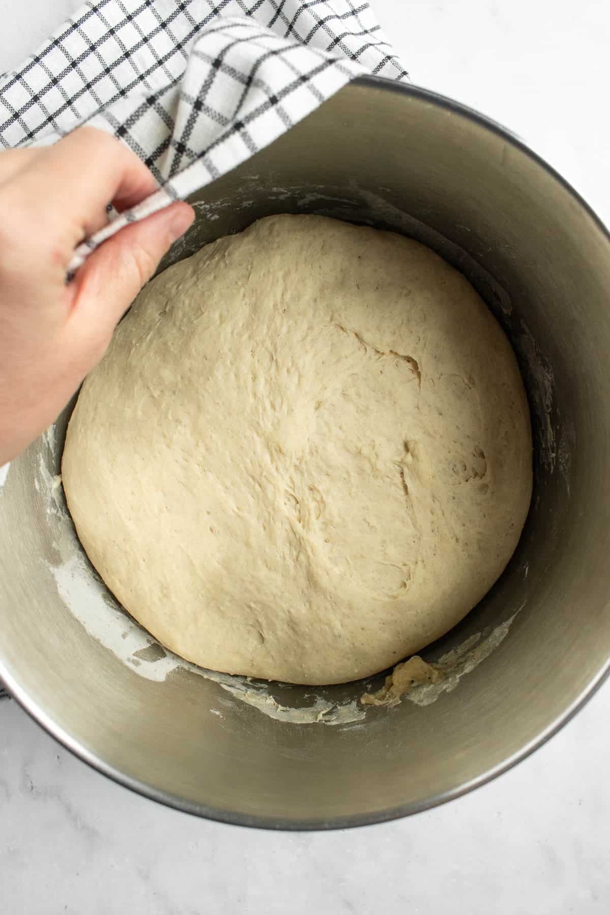 A large risen dough in a metal bowl partially covered by a black and white linen.