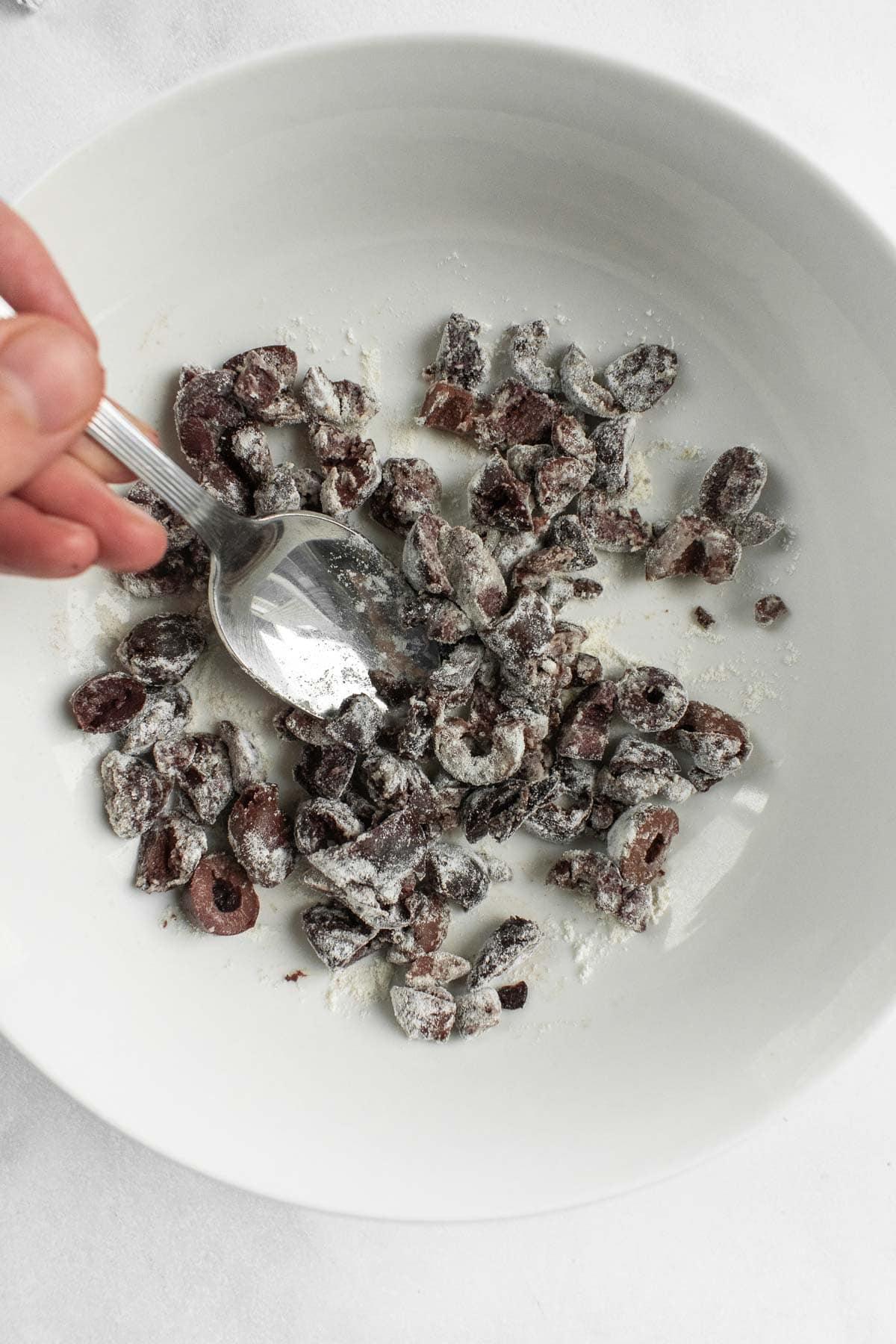A hand mixing chopped olives in flour with a spoon in a bowl.