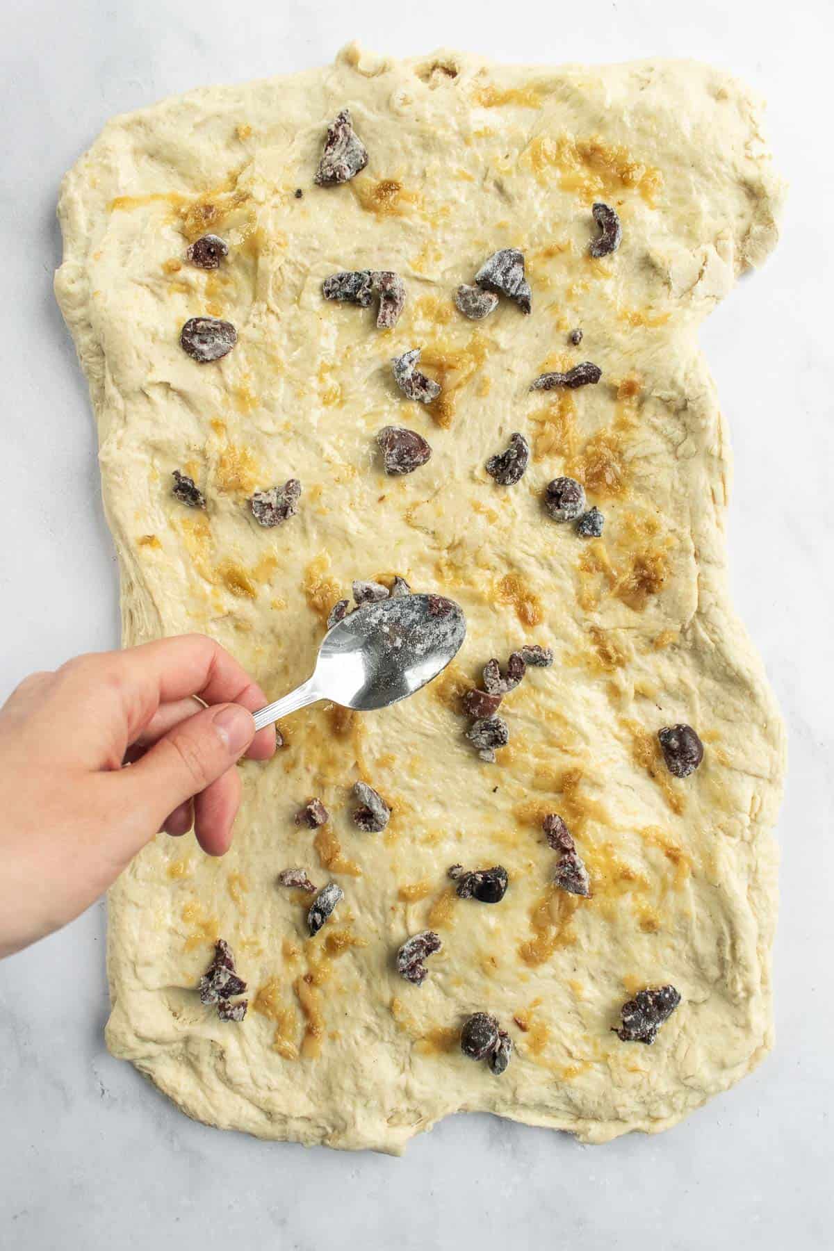 A hand spreading out garlic paste and chopped olives with a spoon onto bread dough.