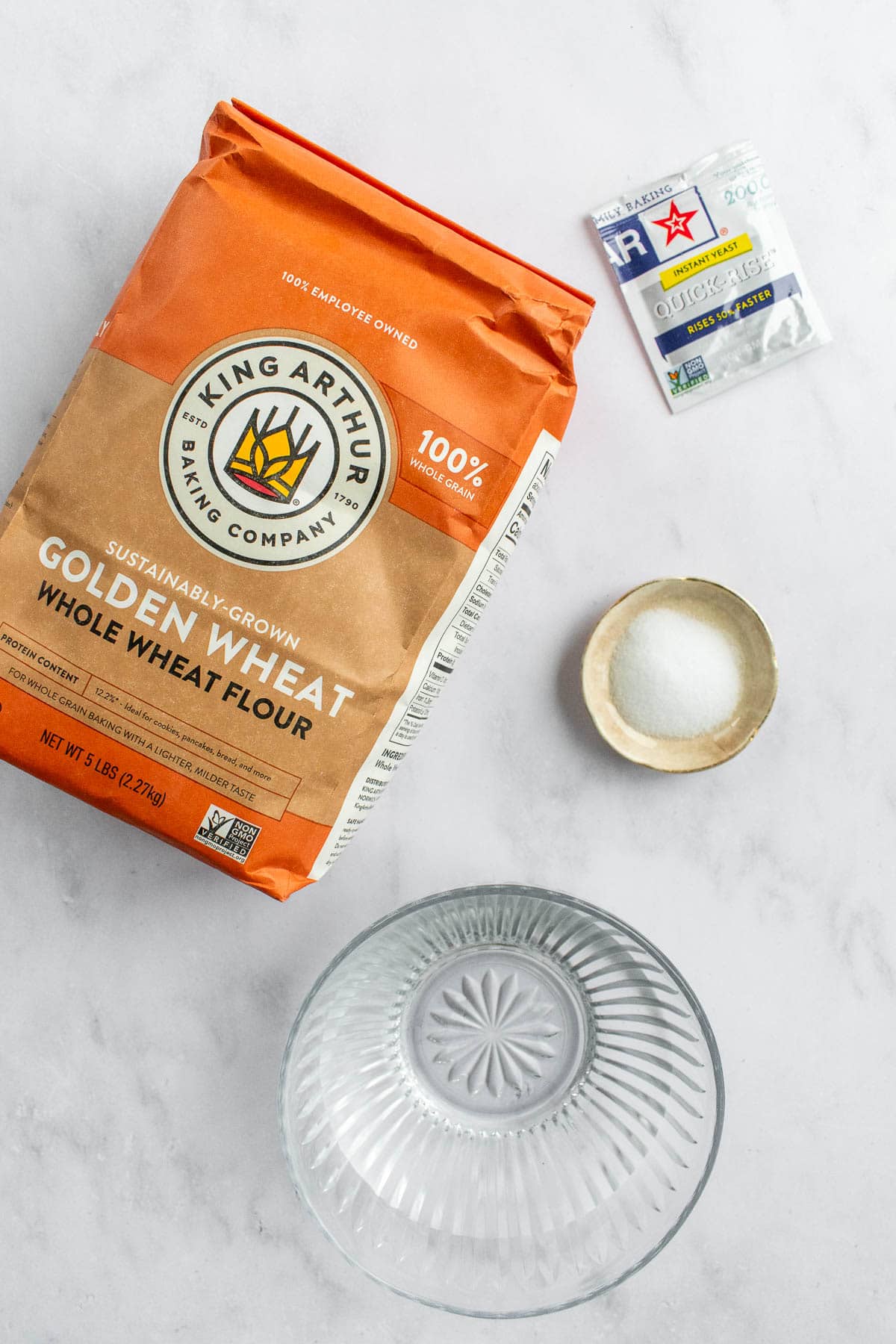 A bag of King Arthur golden wheat flour next to a bowl of water, a dish of salt, and a packet of yeast.