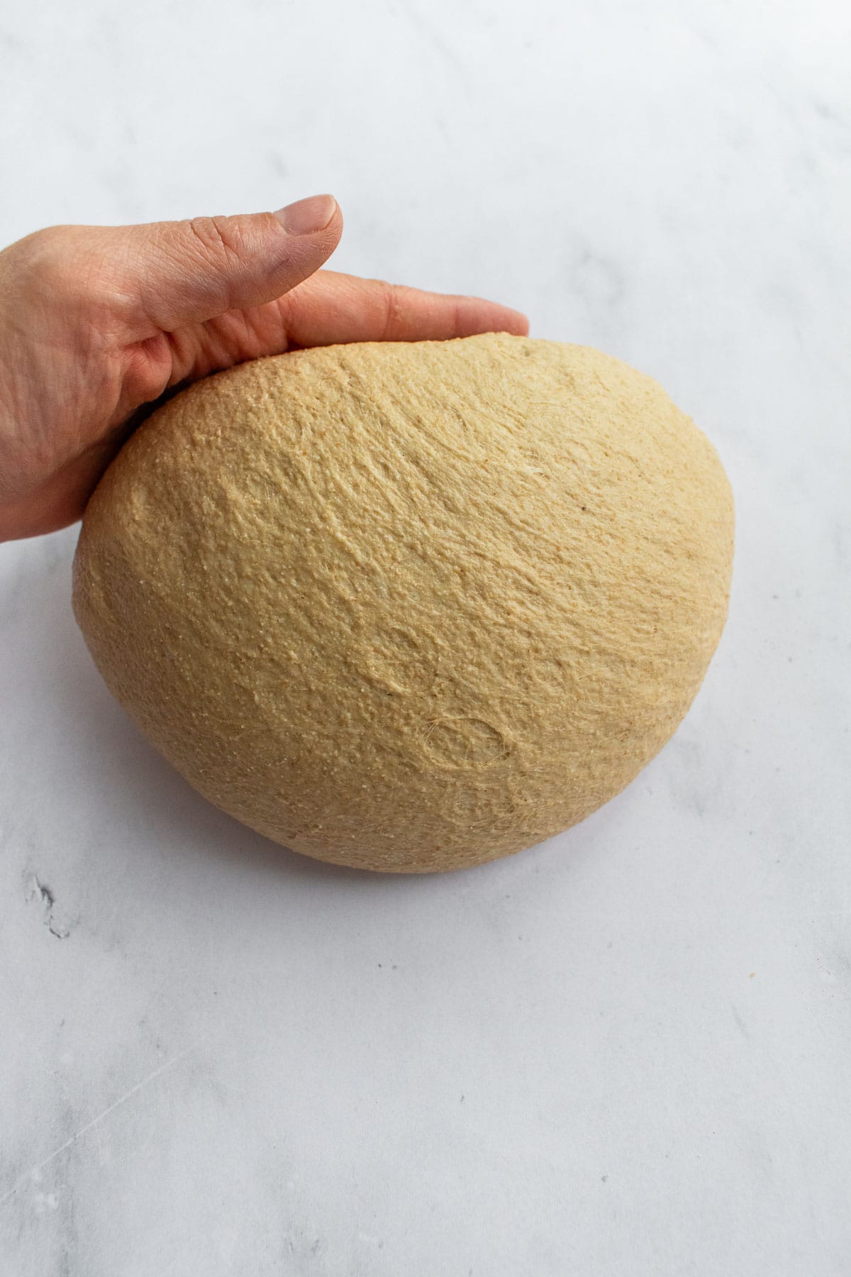 A hand shaping light beige bread dough on a marble surface.