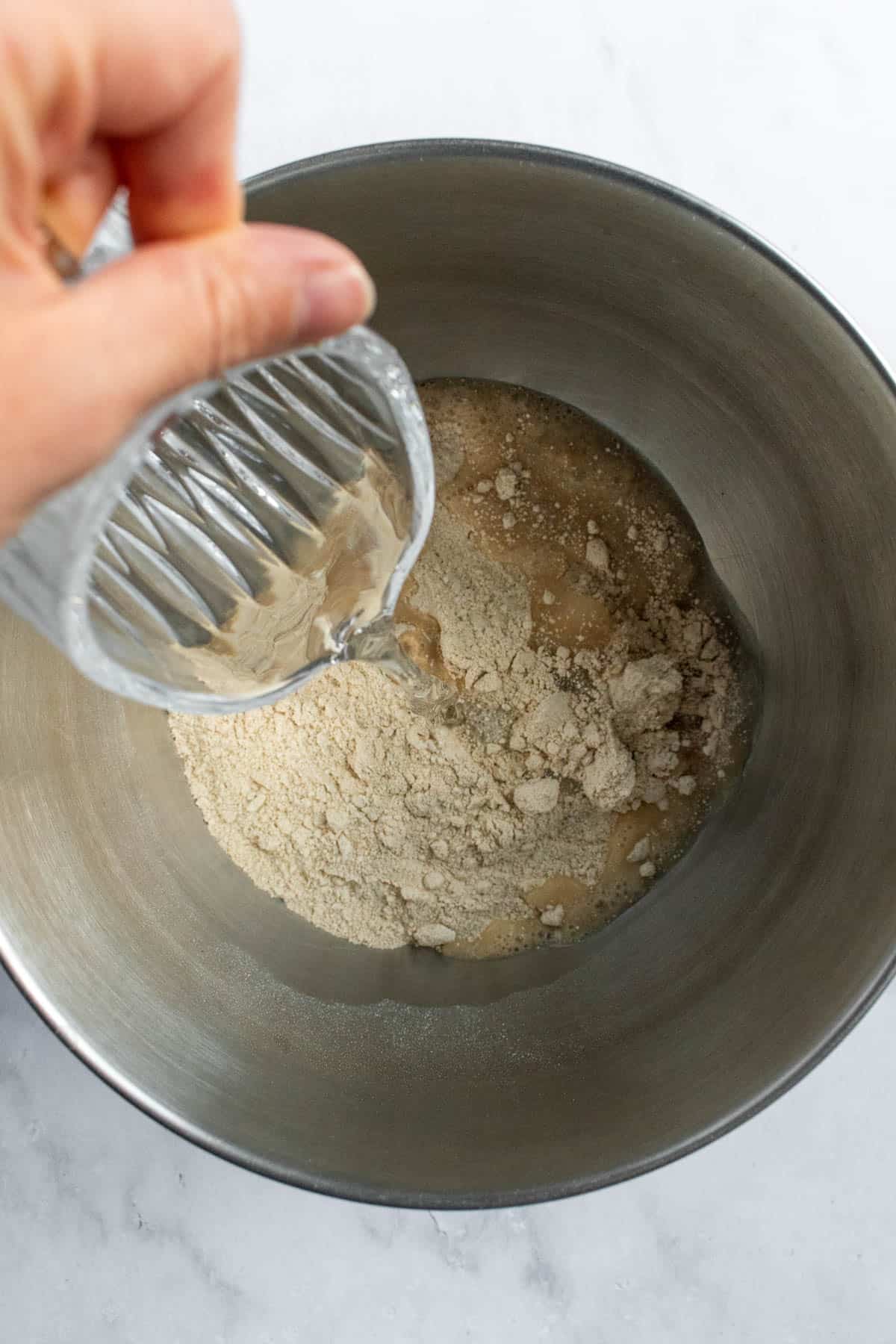 A hand pouring water into a metal bowl of white wheat flour.
