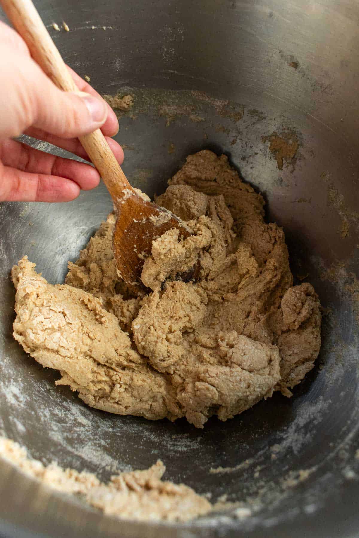 A hand mixing wheat flour and water in a metal bowl with a large wooden spoon.