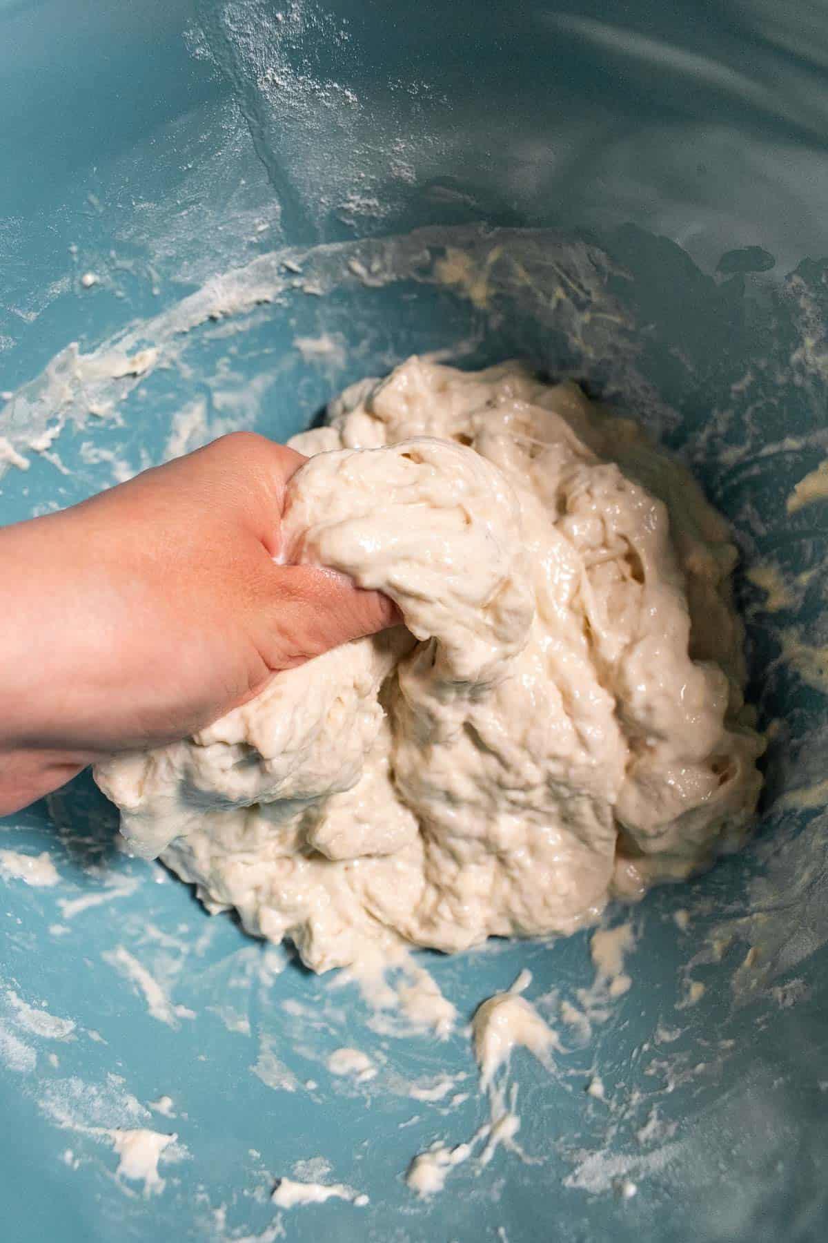 A hand squeezing sticky bread dough in a large blue mixing bowl.
