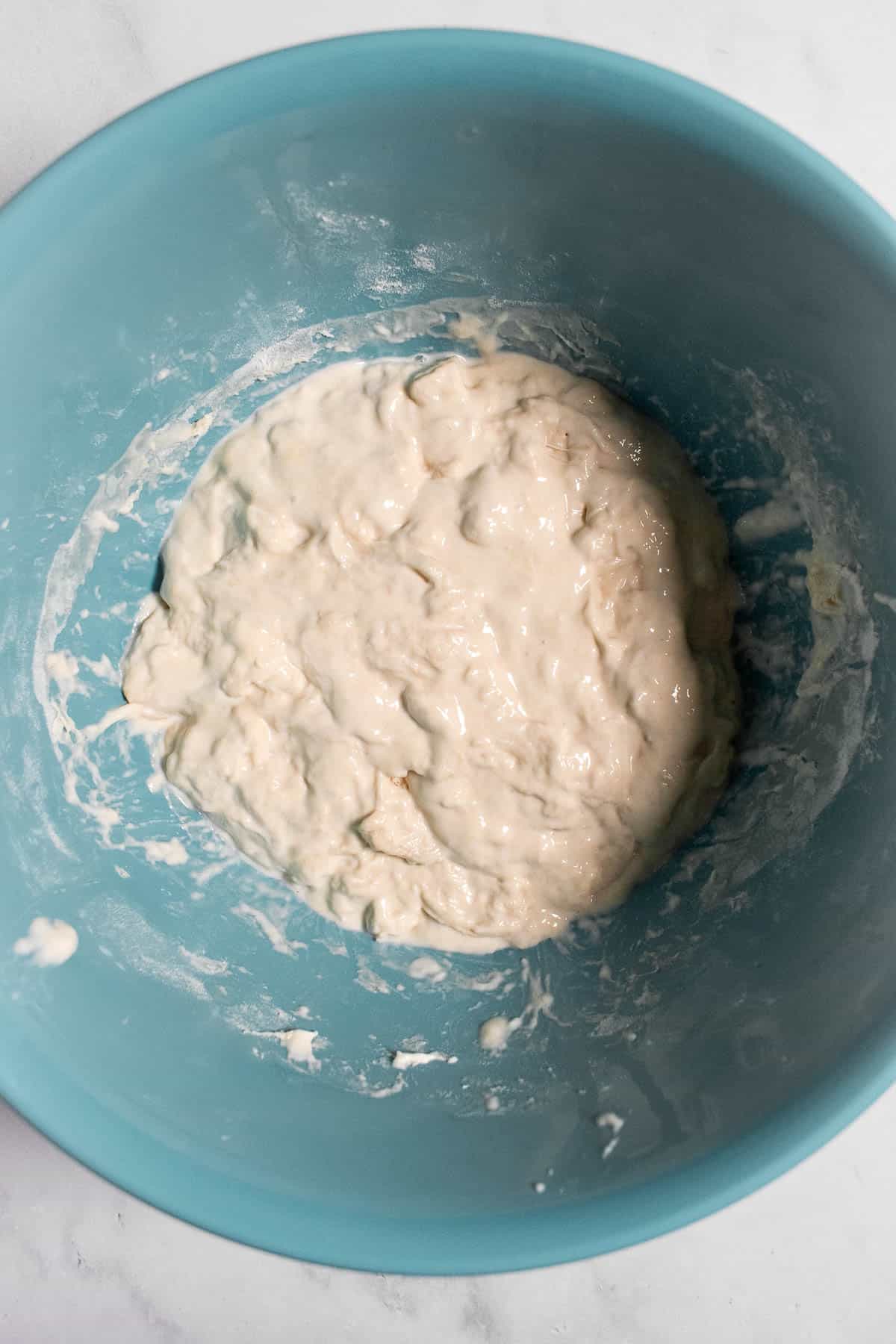 Sticky, white bread dough in a blue mixing bowl on a light surface.