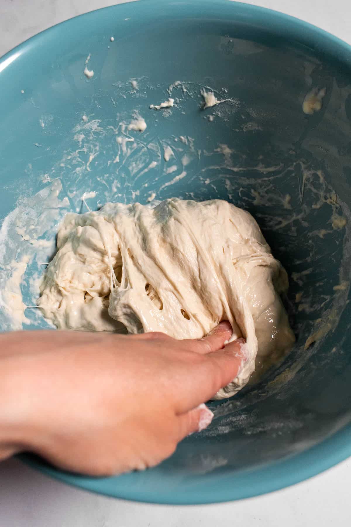 A hand folding bread dough over itself in a blue mixing bowl.