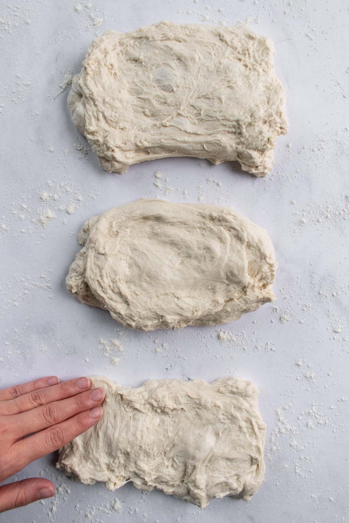 A hand pressing out a rectangle of baguette dough next to two more pieces of dough.