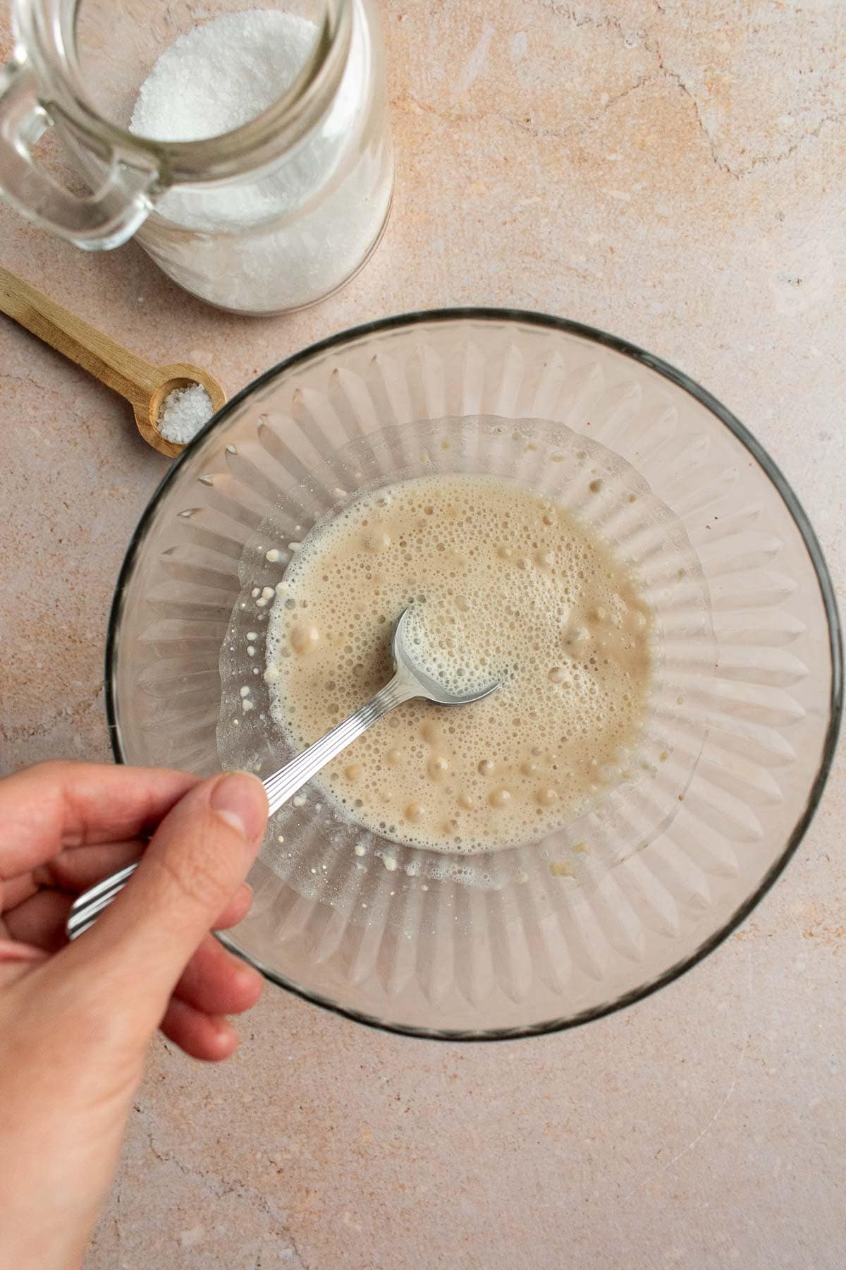 A hand mixing frothy yeast and water next to a glass jar of kosher salt.