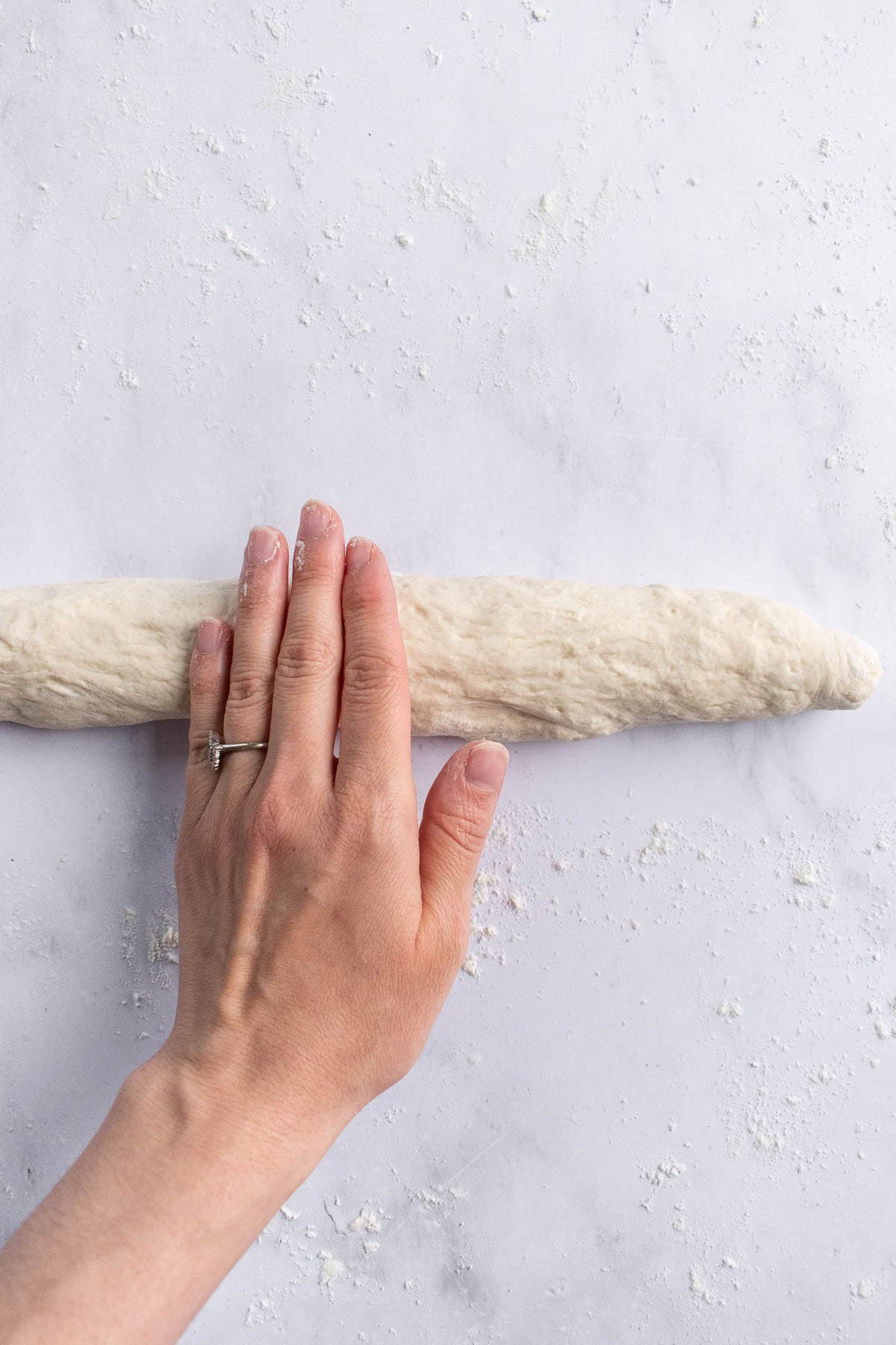 A hand rolling out a loaf of baguette dough with tapered ends.