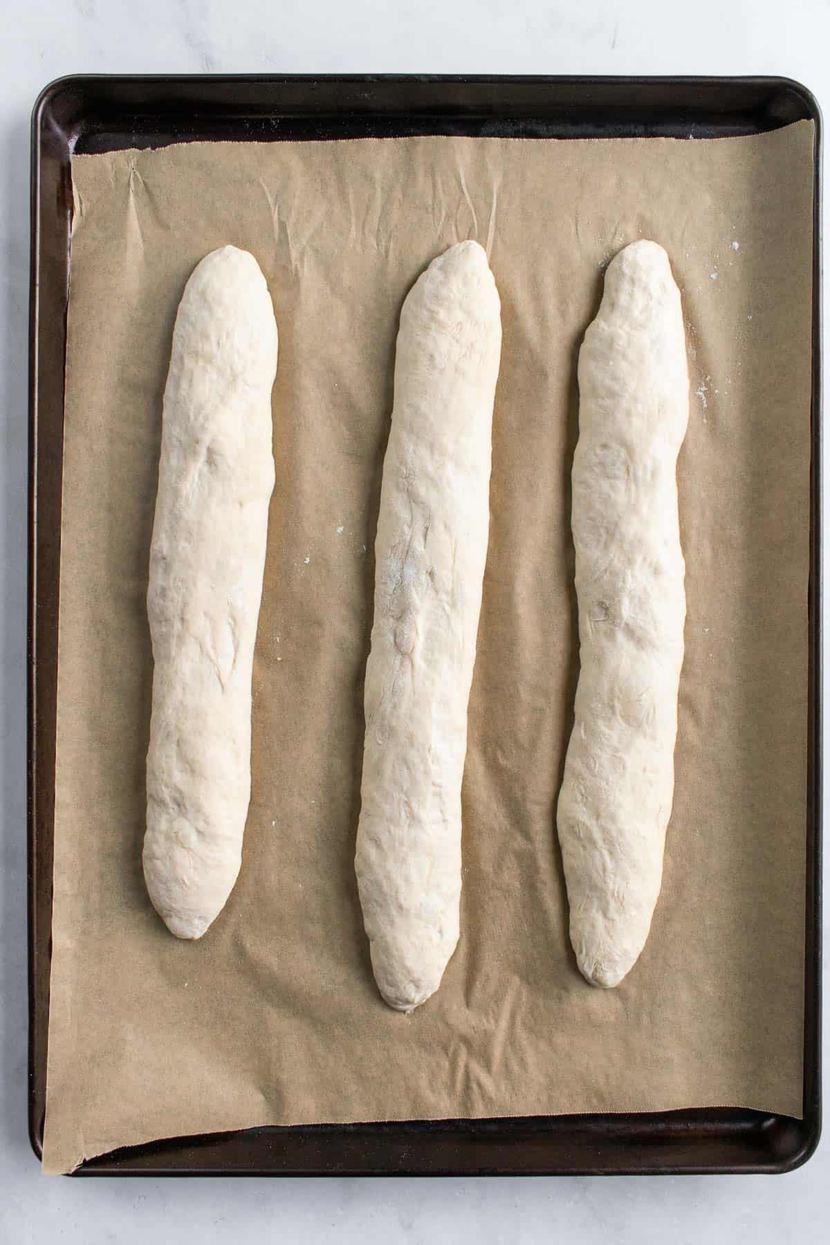 Three unbaked loaves of baguette bread on a baking sheet lined with parchment paper.
