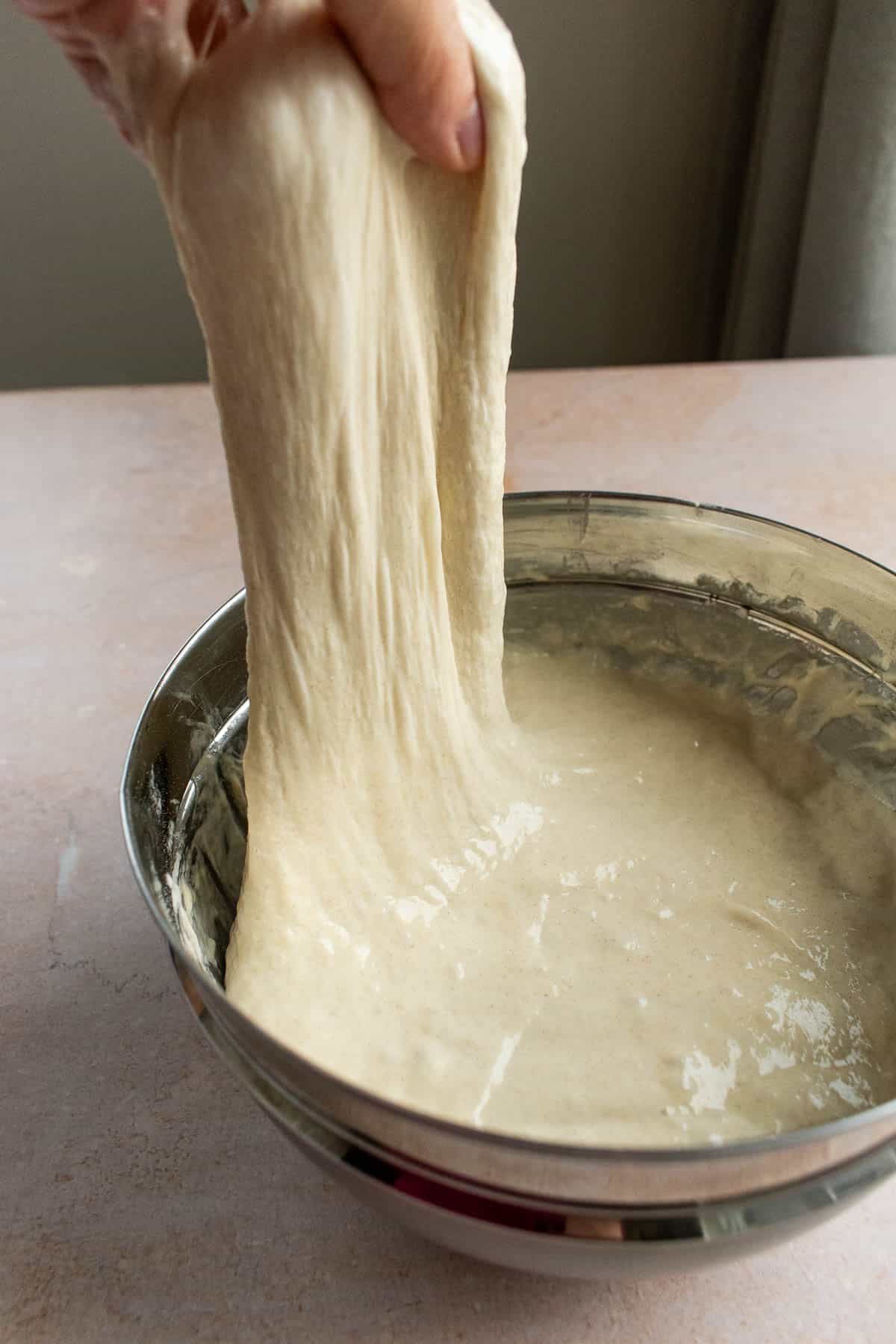 A hand pulling sticky, wet dough out of a metal mixing bowl.