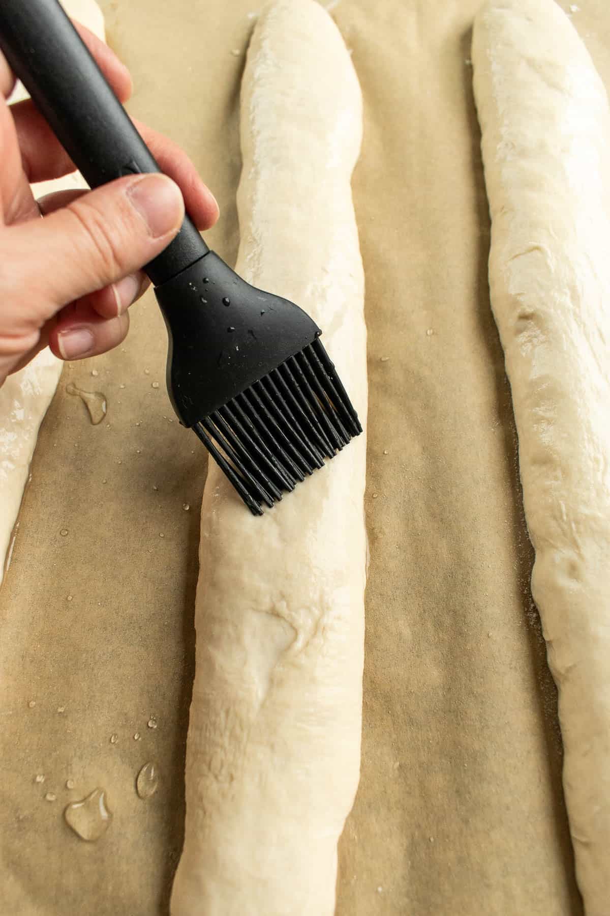 A hand brushing water onto bread dough loaves with a black pastry brush.