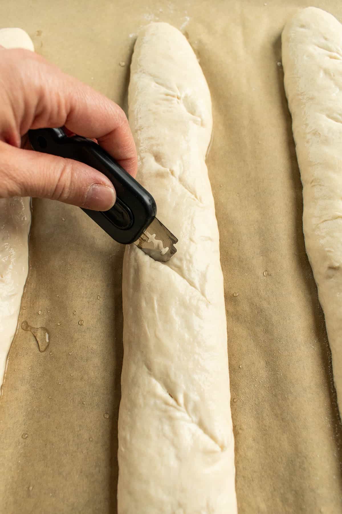 A hand scoring baguette dough with a sharp bread lame.