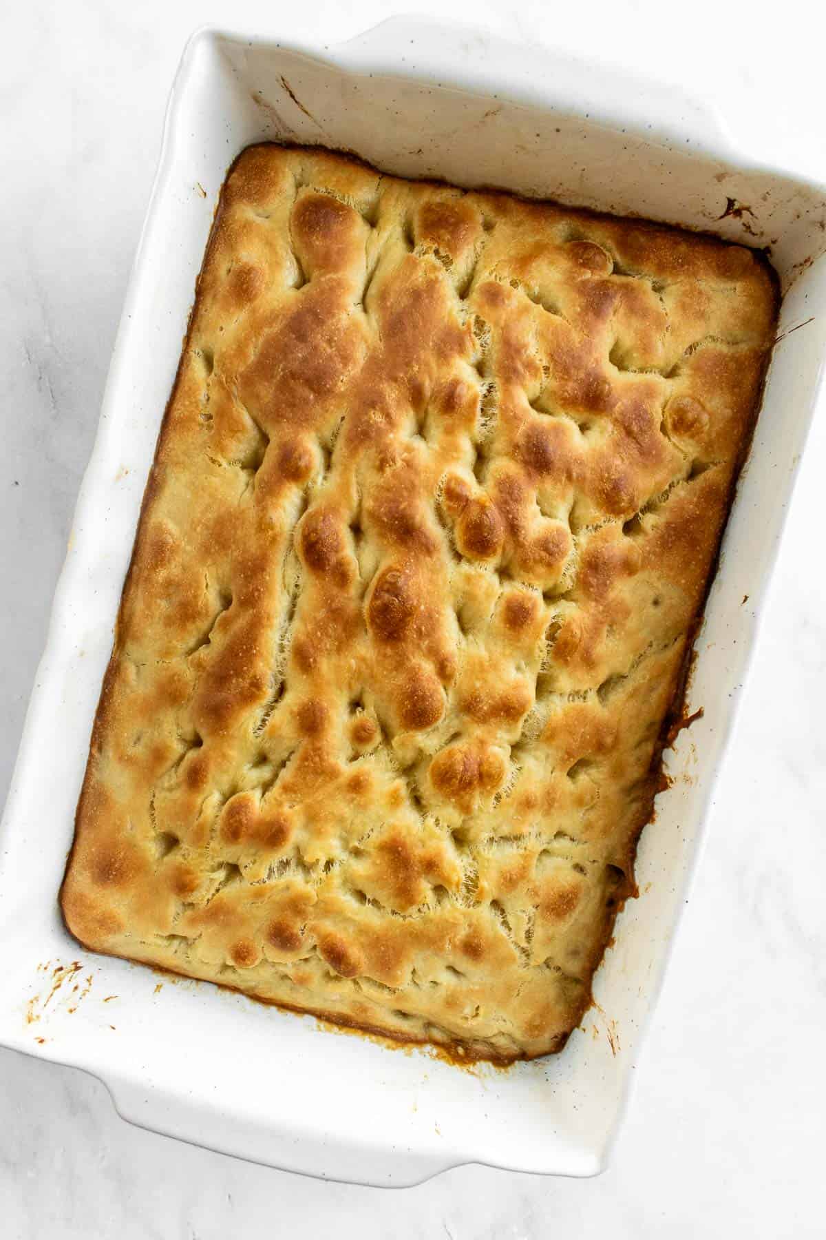 Baked golden focaccia bread in a white baking dish on a light surface.