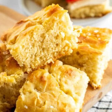 Squares of fluffy focaccia bread on a wooden board, next to a plate of two sandwiches.