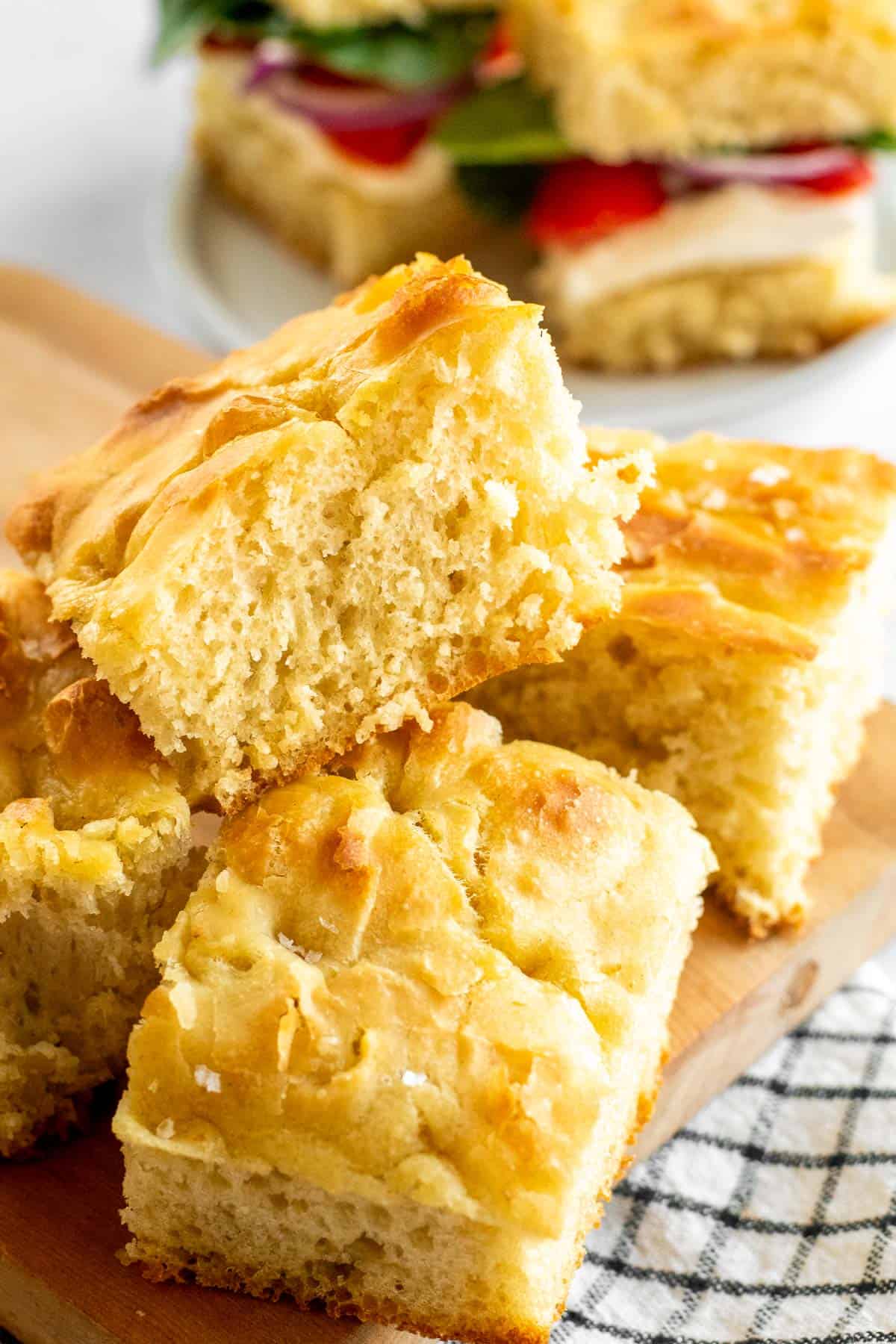 Squares of fluffy focaccia bread on a wooden board, next to a plate of two sandwiches. 