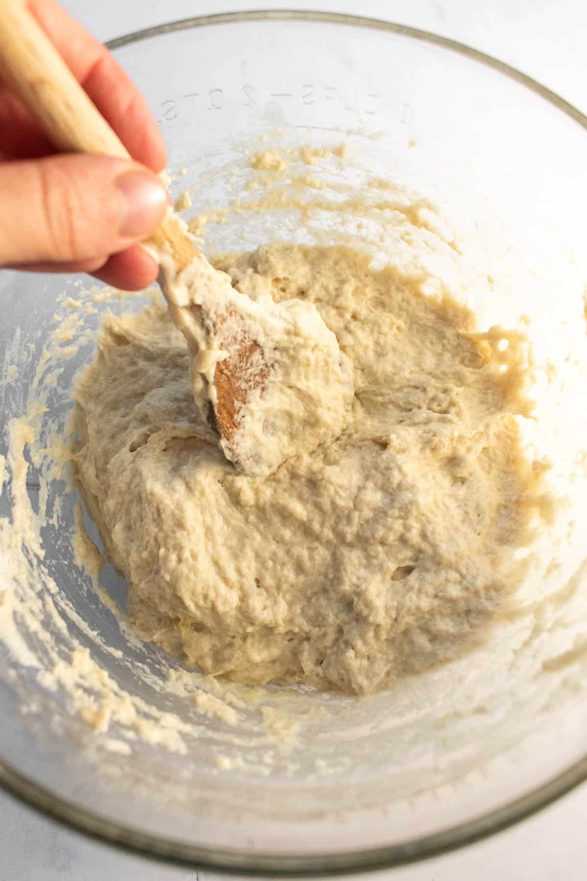 A hand mixing wet dough with a wooden spoon in a glass bowl.