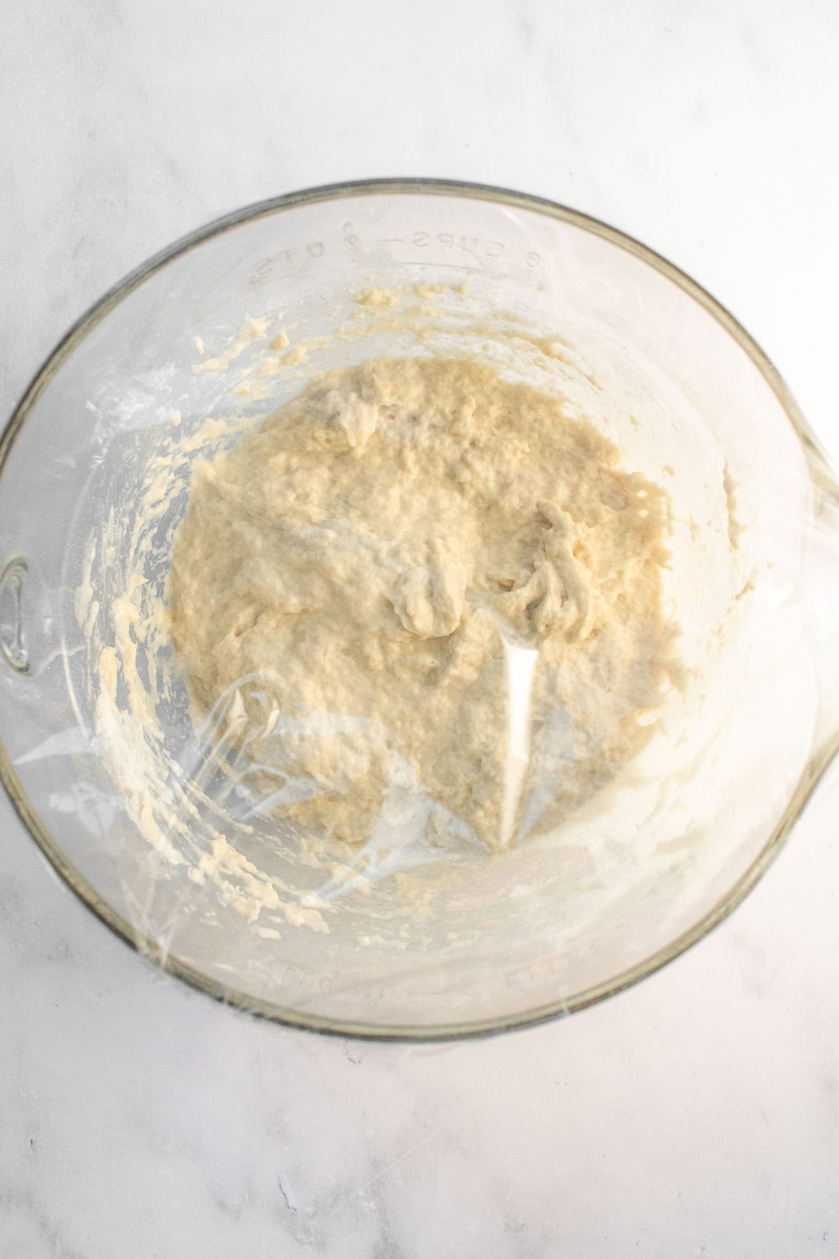 A glass bowl filled with wet focaccia bread dough covered with plastic wrap.