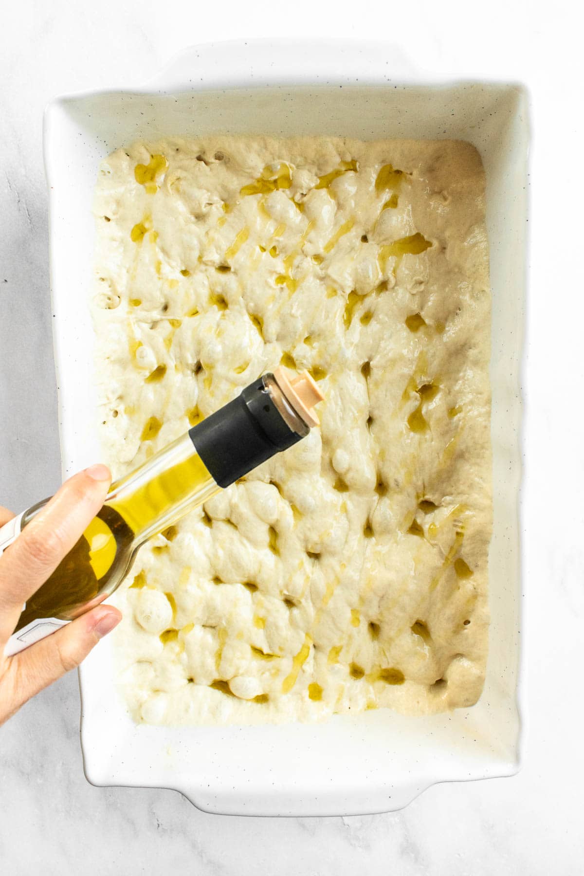 A hand pouring olive oil from a bottle onto dimpled focaccia dough in a white baking dish.