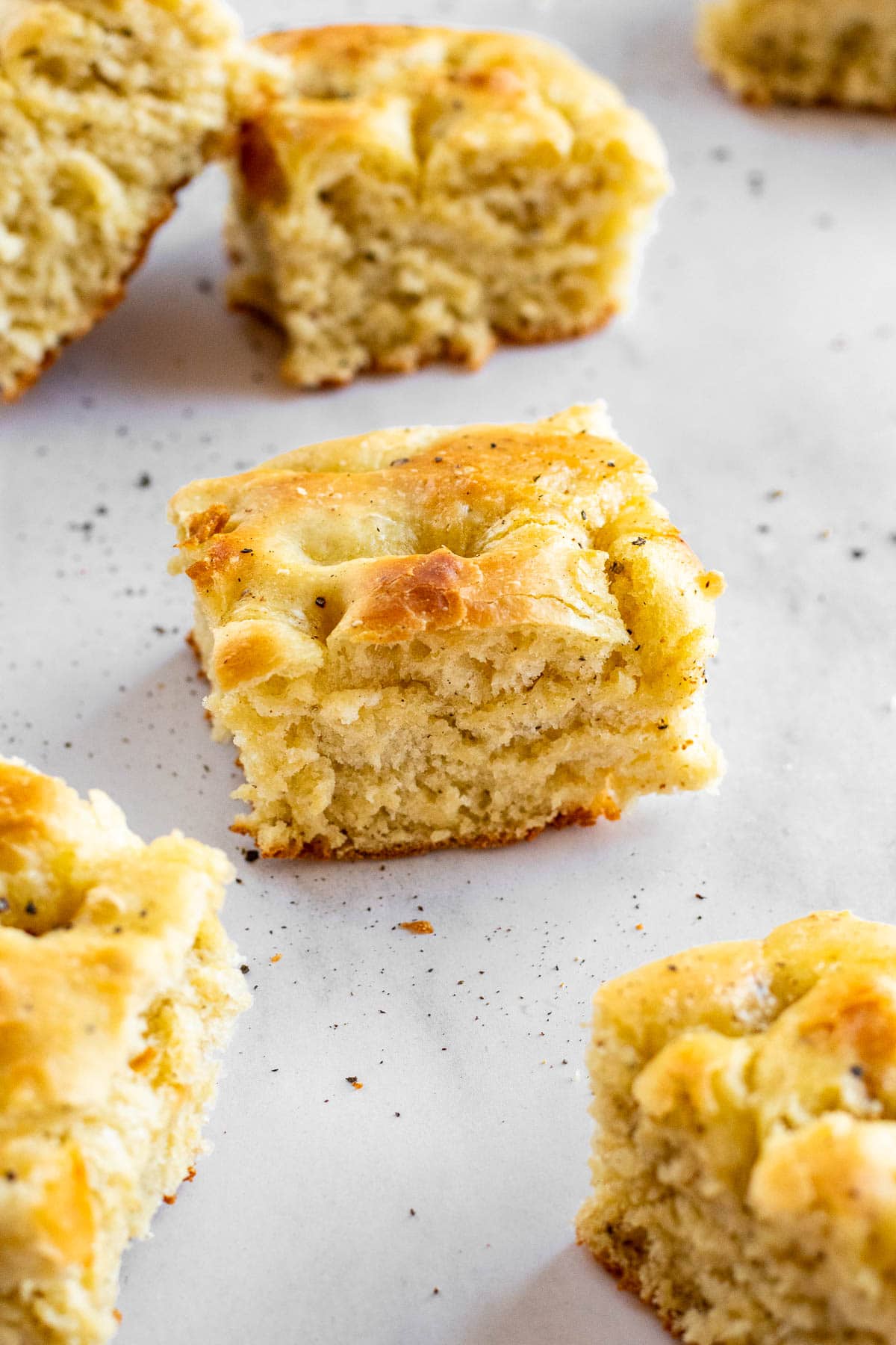 Half a dozen squares of golden focaccia topped with black pepper spread out on a light surface.